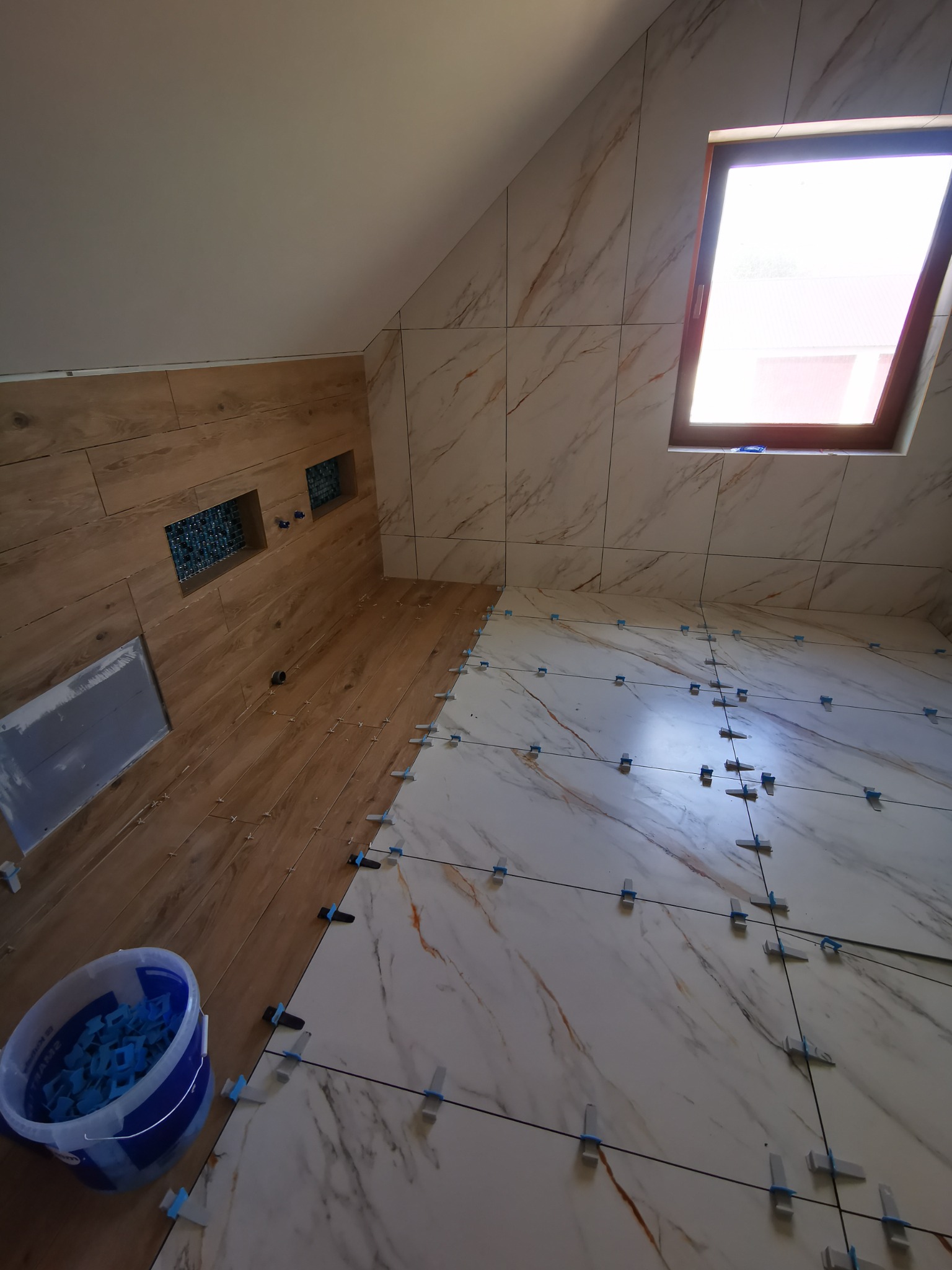 Łazienka na poddaszu w trakcie układania płytek imitujących marmur na podłodze i ścianach z oknem dachowym; widoczne krzyżyki dystansowe i wiadro z klinami do płytek.