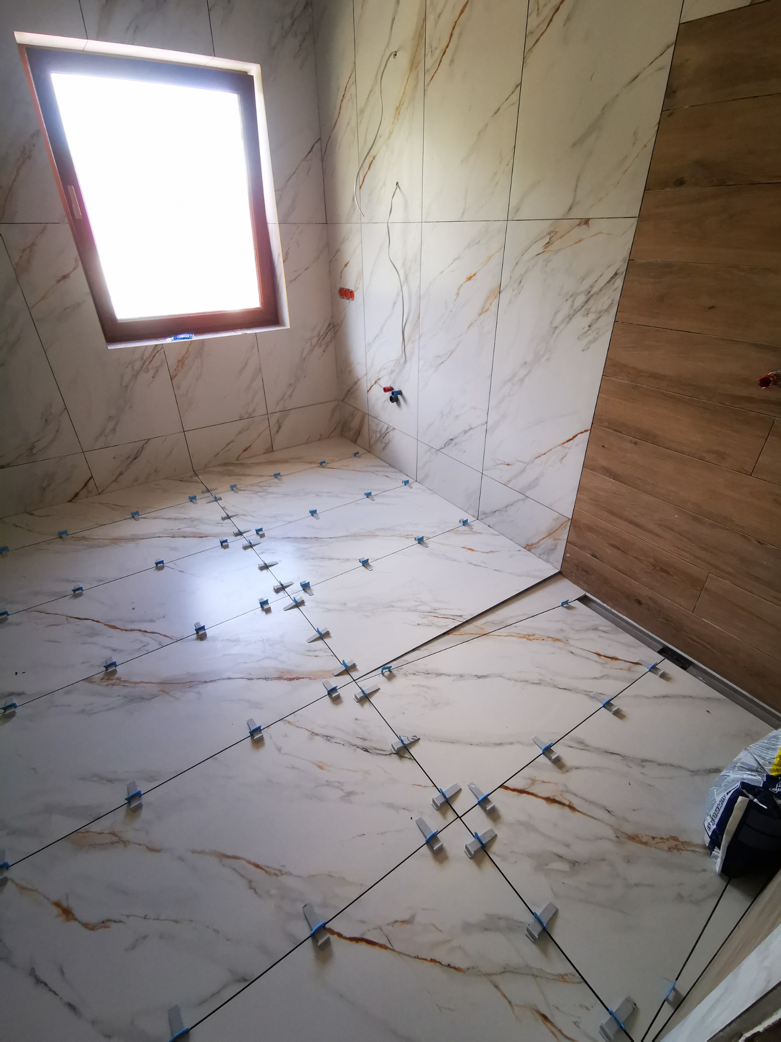 Białe marmurowe płytki podłogowe i ścienne z brązowymi żyłami, ułożone w łazience z oknem dachowym, z użyciem klipsów poziomujących.