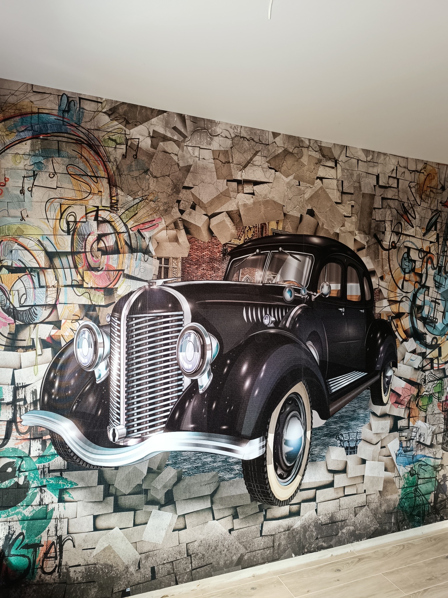 Fototapeta na ścianie przedstawiająca czarny, klasyczny samochód w stylu lat 30. przebijający się przez mur z cegieł, z elementami graffiti i nutami muzycznymi w tle.