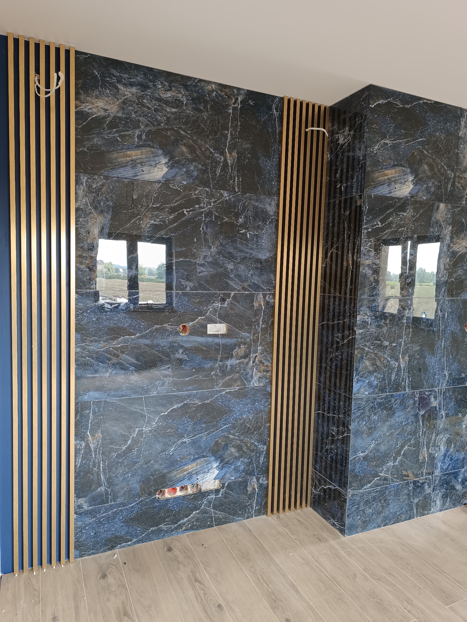 Ściana wyłożona dużymi płytami imitującymi ciemny marmur z białymi żyłami, z drewnianymi lamelami po bokach, widoczne niedokończone gniazdka elektryczne i fragment parkietu.