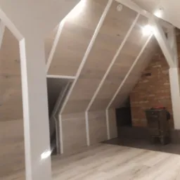 Wykończone poddasze z drewnianą boazerią na skosach, białe listwy wykończeniowe, białe filary i fragment ściany z czerwonej cegły z piecem.