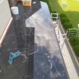 Błyszczące, czarne płytki imitujące marmur na balkonie, częściowo zakryte niebieską ścierką, z widocznym odbiciem nieba i drabiny, oraz fragmentem ogrodu z zielonym trawnikiem i krzewami.