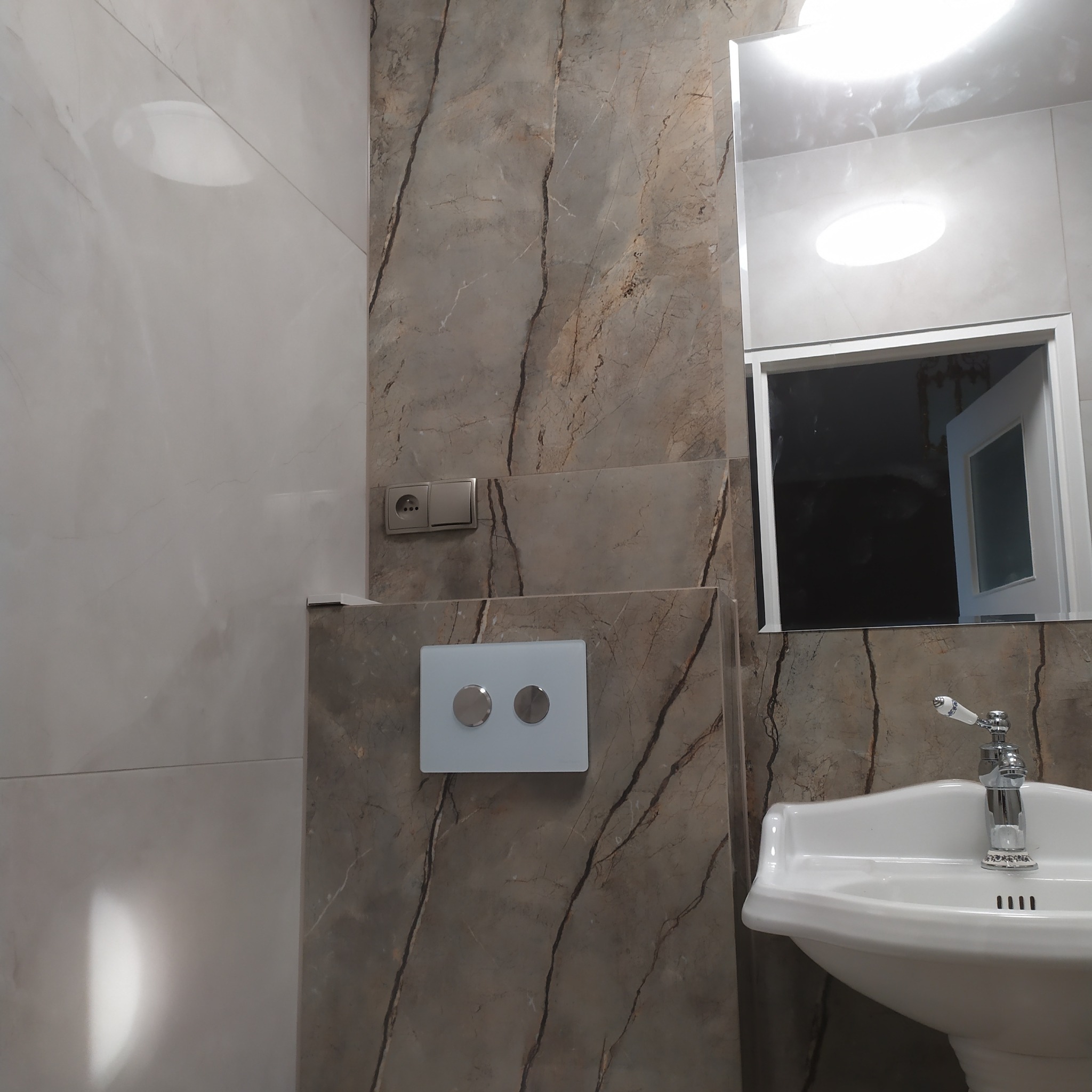 Umywalka z ozdobną baterią w wykończonej łazience, ściany wyłożone płytkami imitującymi marmur, widoczny przycisk spłuczki i gniazdko elektryczne.