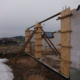 Daniel Gut USŁUGI OGÓLNOBUDOWLANE - Budowa domu w trakcie wznoszenia ścian: widoczne szalunki z płyt OSB, podpory z bali oraz fragmenty muru z betonu komórkowego, teren budowy z resztkami śniegu.