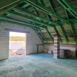 Daniel Gut USŁUGI OGÓLNOBUDOWLANE - Surowy stan poddasza z widoczną konstrukcją dachu pomalowaną na zielono, mury z betonu komórkowego, fragment komina i widok na zewnątrz przez otwór okienny.