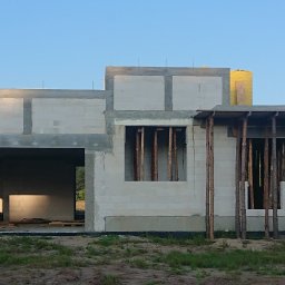 Daniel Gut USŁUGI OGÓLNOBUDOWLANE - Surowy stan budynku z betonu komórkowego w trakcie budowy, widoczne otwory okienne z drewnianymi stemplami, niezadaszone wejście i fragment lasu w tle.