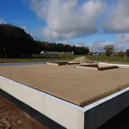 Przygotowany teren pod wylewkę fundamentów z widoczną hydroizolacją pionową oraz szalunkami pod przyszłe ściany nośne, w tle krajobraz wiejski.