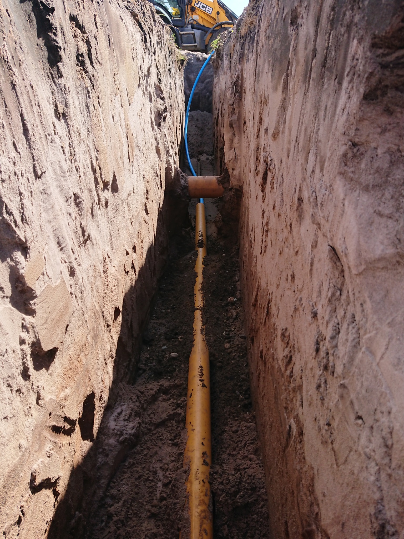 Wykop w ziemi z ułożonymi rurami, widoczny fragment koparki JCB na górze, pionowe ściany wykopu z nierówną strukturą.