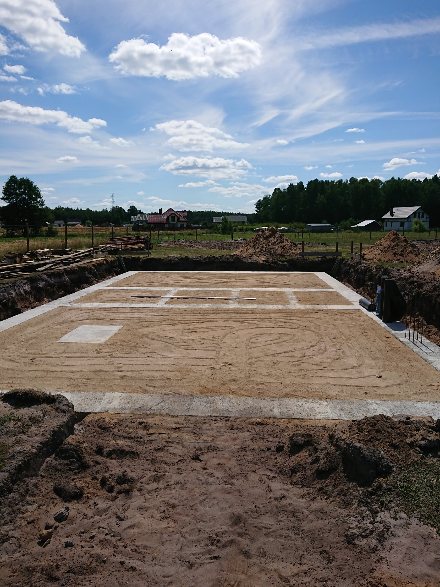Przygotowany teren pod fundamenty z widocznymi wykopami i piaskiem, na horyzoncie zabudowania wiejskie w słoneczny dzień.