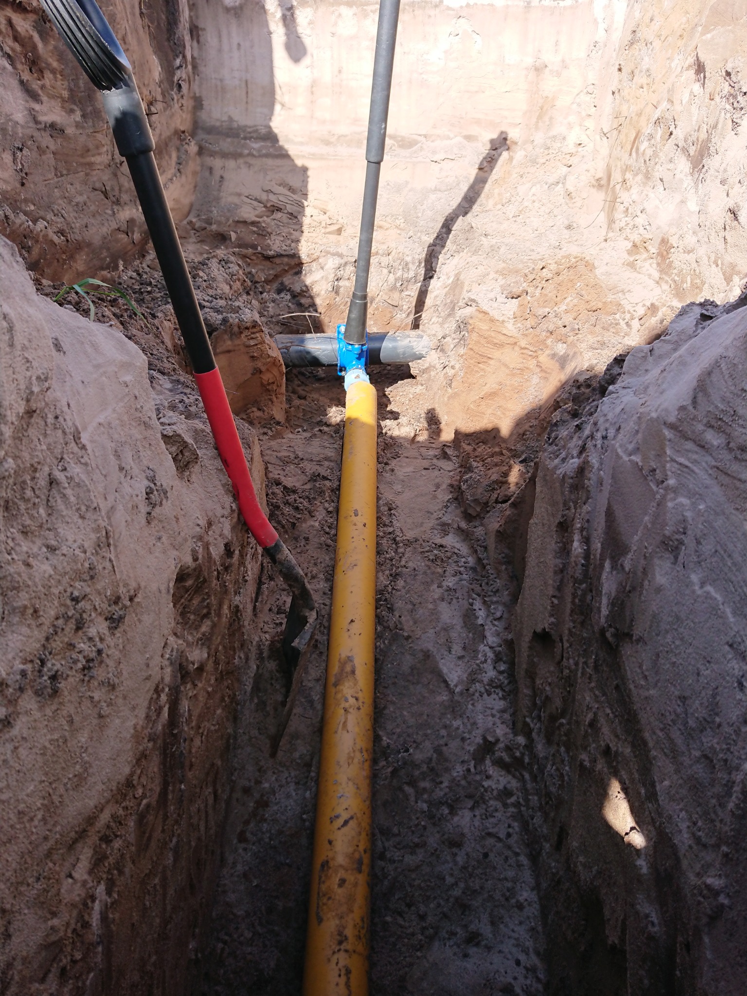Wykop z żółtą rurą gazową połączoną niebieskim złączem, widoczna łopata z czerwoną rączką i cień osoby stojącej nad wykopem.