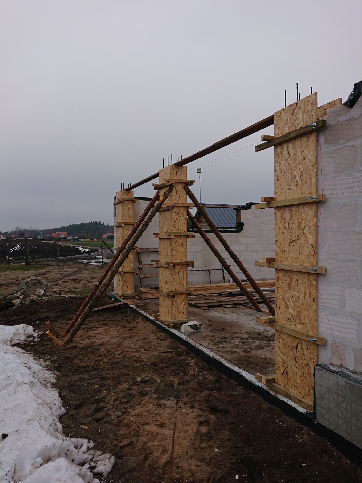 Budowa domu w trakcie wznoszenia ścian: widoczne szalunki z płyt OSB, podpory z bali oraz fragmenty muru z betonu komórkowego, teren budowy z resztkami śniegu.