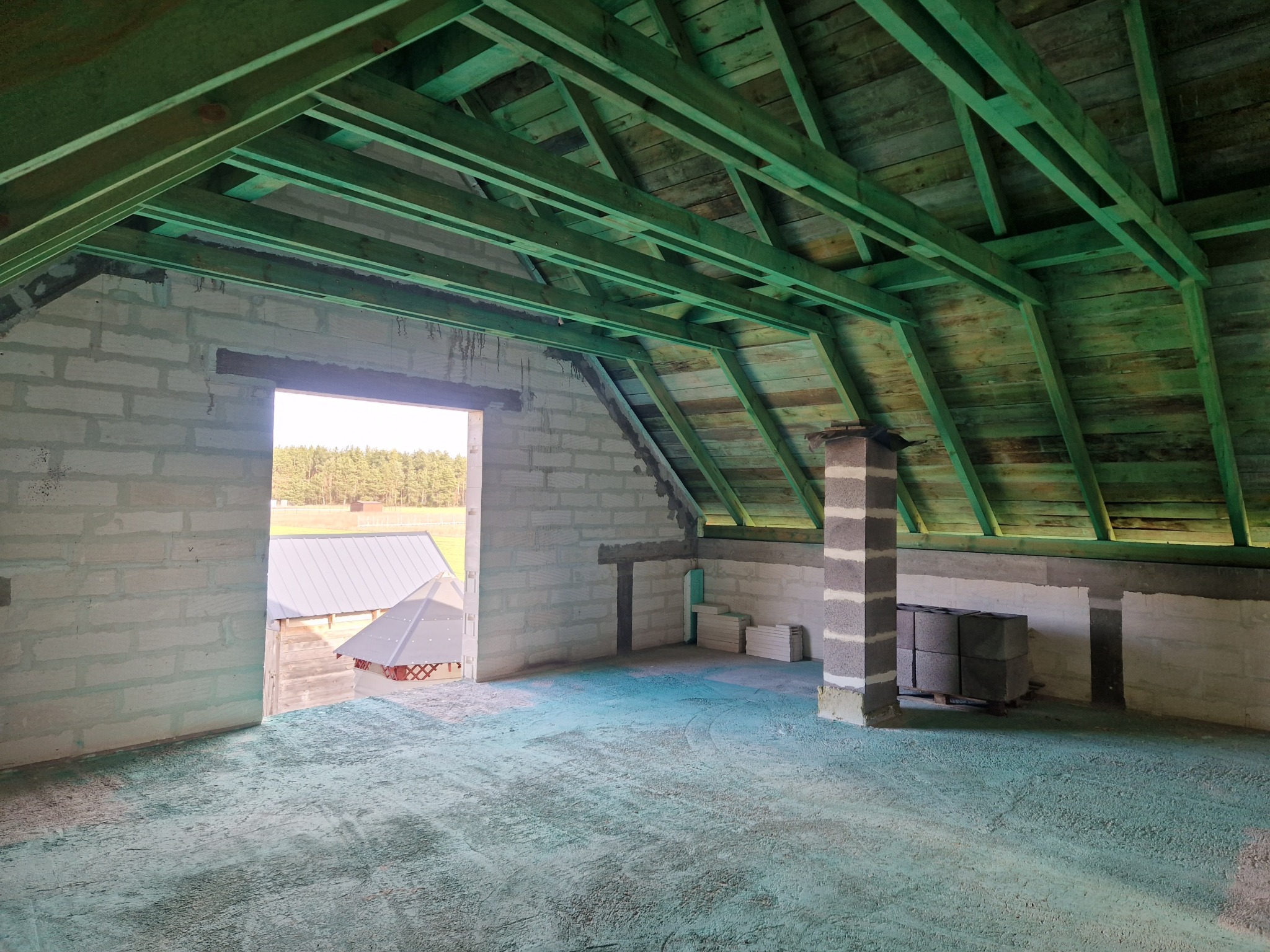 Surowy stan poddasza z widoczną konstrukcją dachu pomalowaną na zielono, mury z betonu komórkowego, fragment komina i widok na zewnątrz przez otwór okienny.