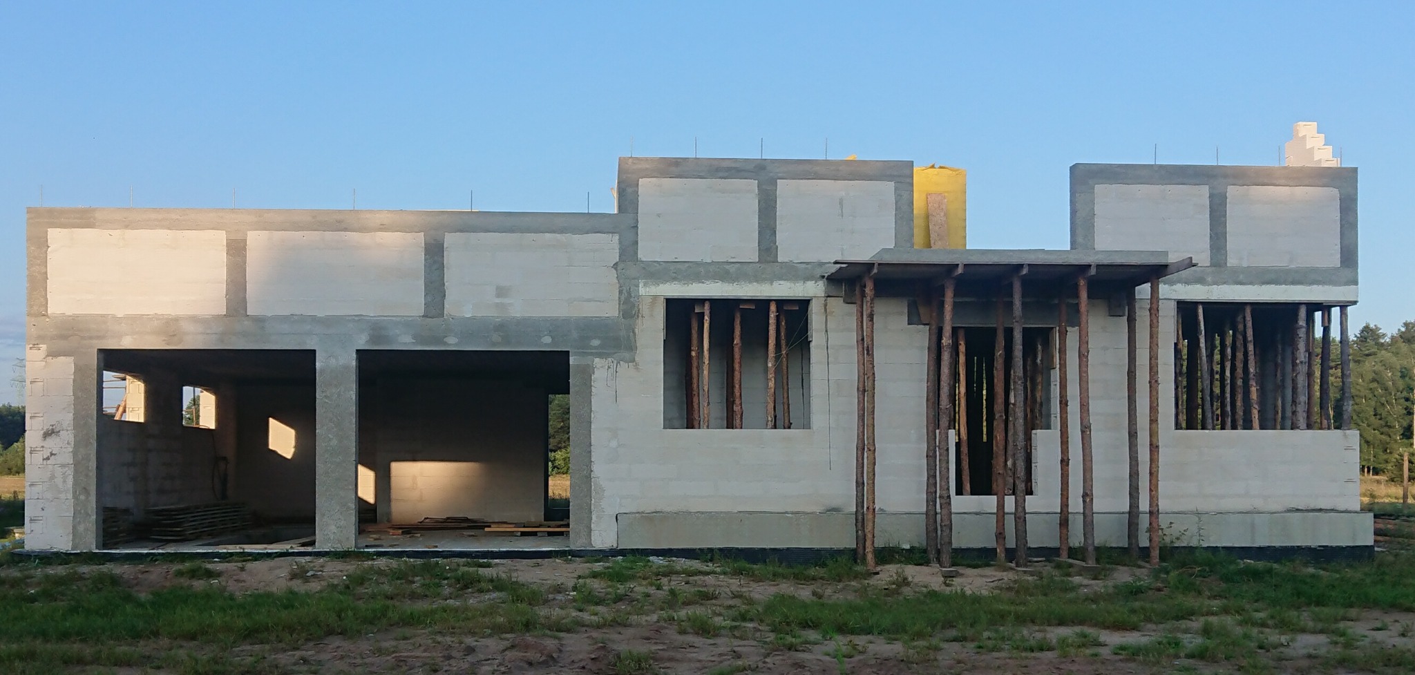 Surowy stan budynku z betonu komórkowego w trakcie budowy, widoczne otwory okienne z drewnianymi stemplami, niezadaszone wejście i fragment lasu w tle.