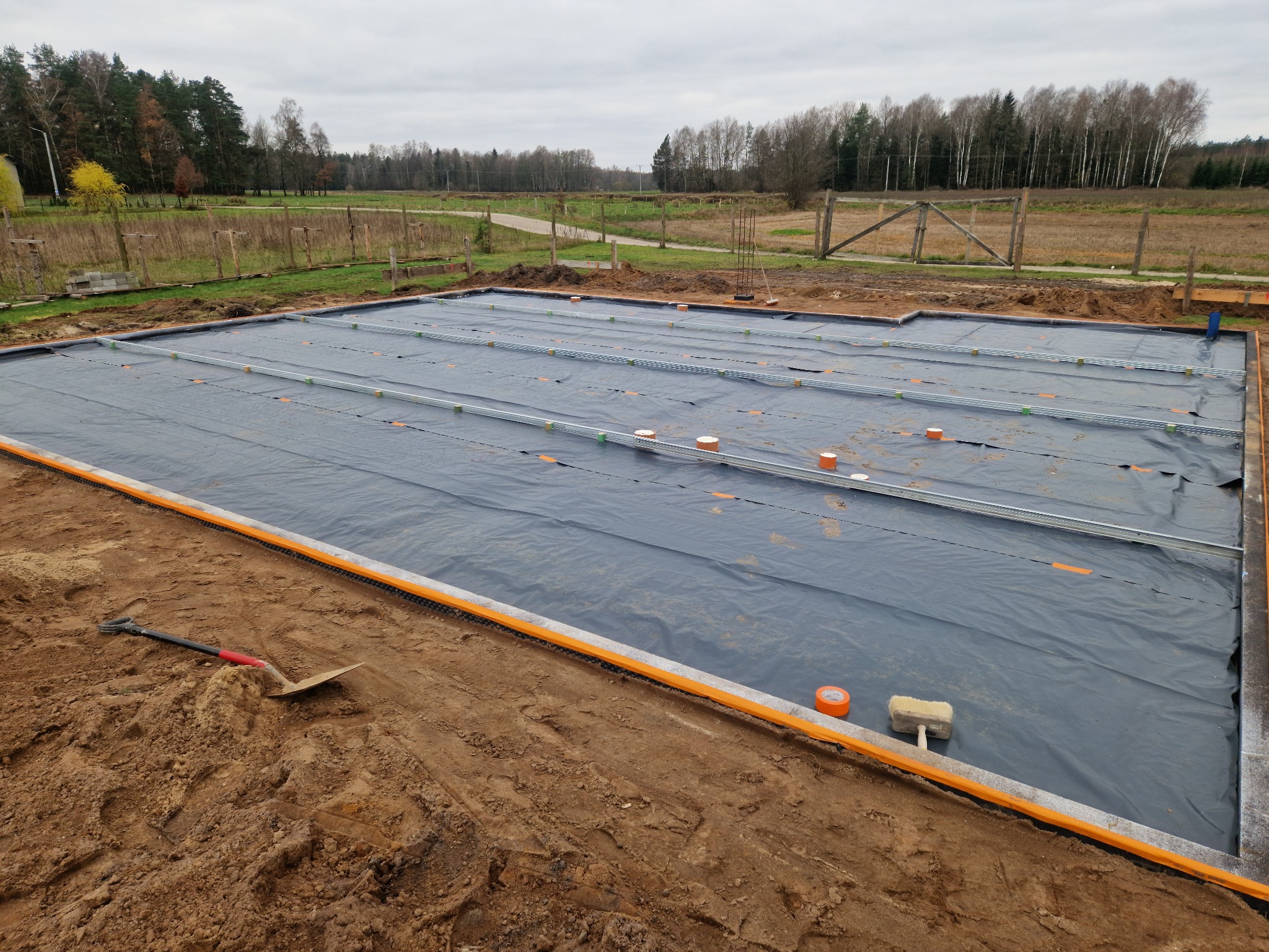 Przygotowany teren pod budowę domu z rozłożoną czarną folią izolacyjną, widoczne metalowe profile, pomarańczowe rury wentylacyjne i taśma, łopata na ziemi, w tle drzewa i brama.