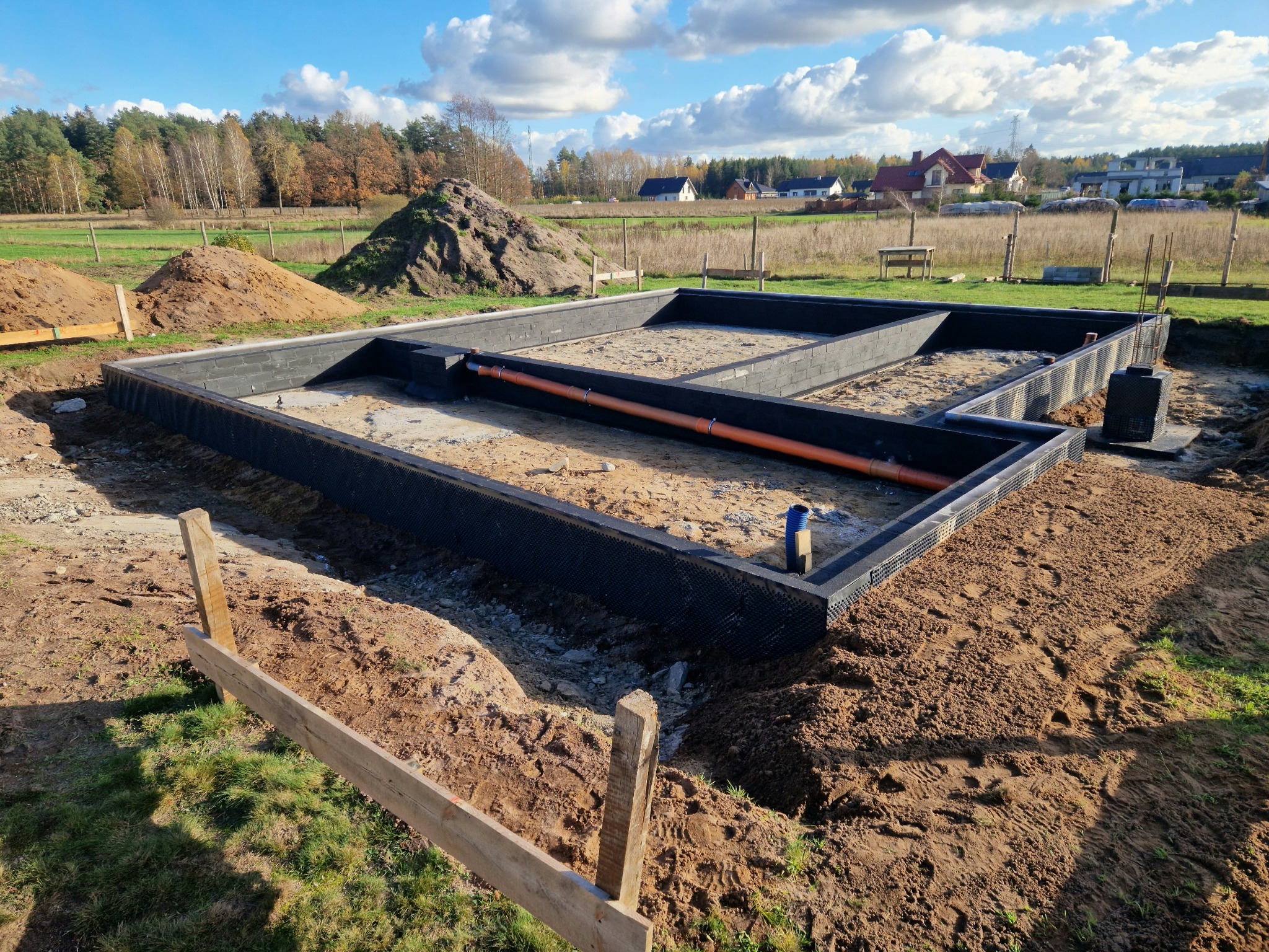 Przygotowany zbrojony fundament pod budowę domu jednorodzinnego z widoczną izolacją przeciwwilgociową oraz rurą kanalizacyjną, na tle krajobrazu z drzewami i zabudowaniami.