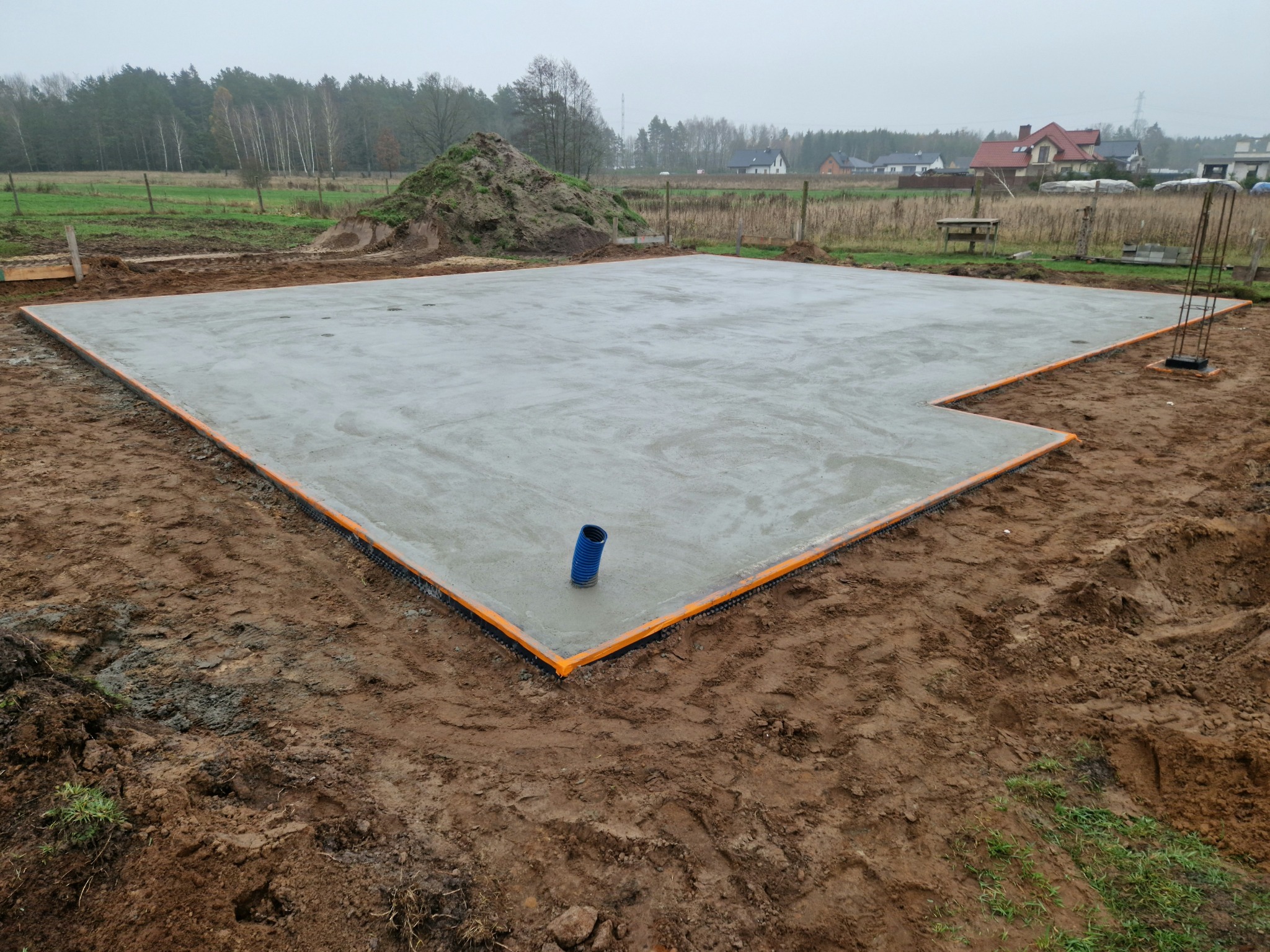 Wylany fundament z widoczną izolacją pionową w postaci pomarańczowej folii, na pierwszym planie wystająca niebieska rura kanalizacyjna, w tle hałda ziemi i zabudowa jednorodzinna.