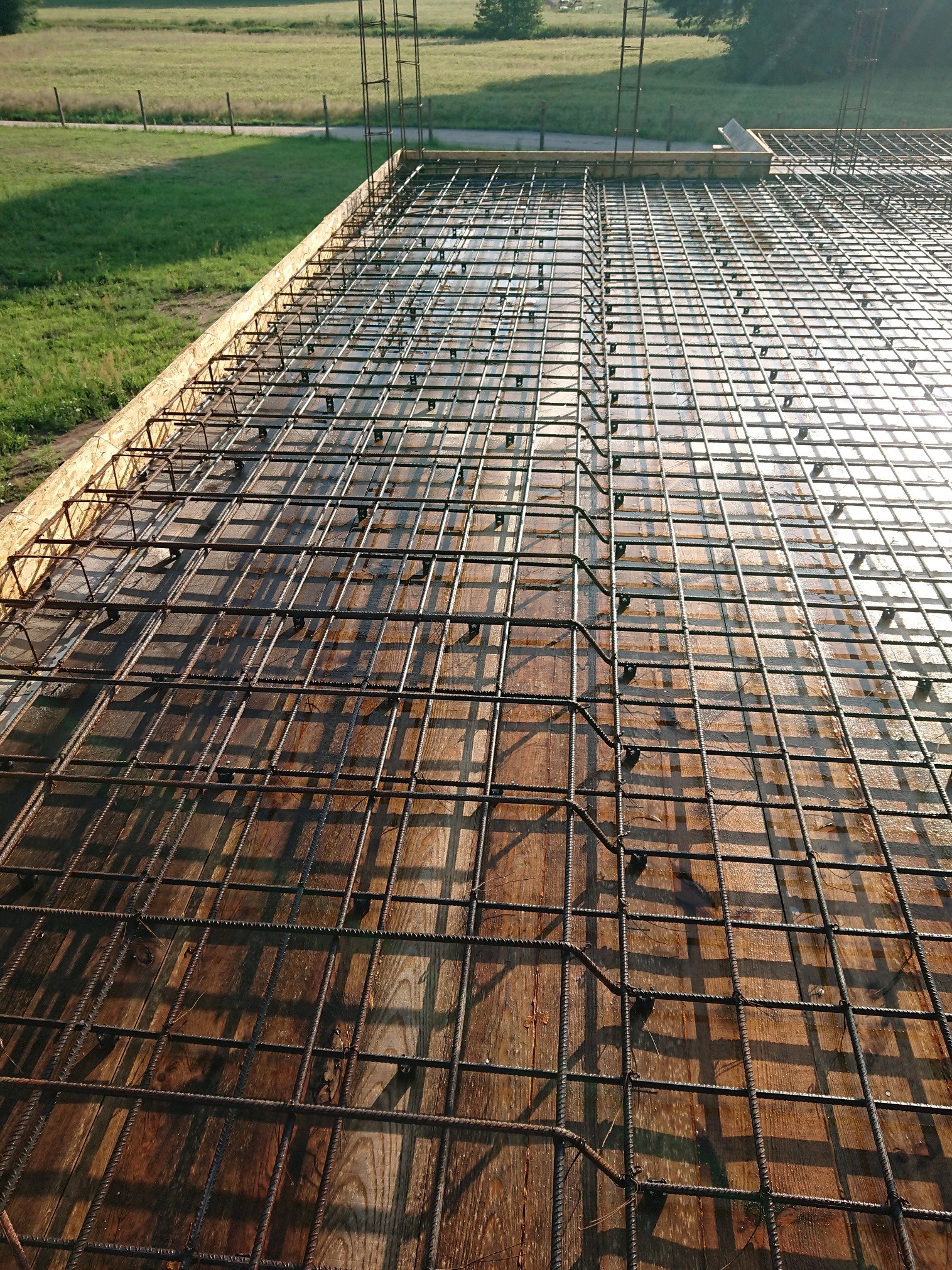 Zbrojenie płyty fundamentowej na drewnianym szalunku, widok z góry, z zielonym trawnikiem w tle.