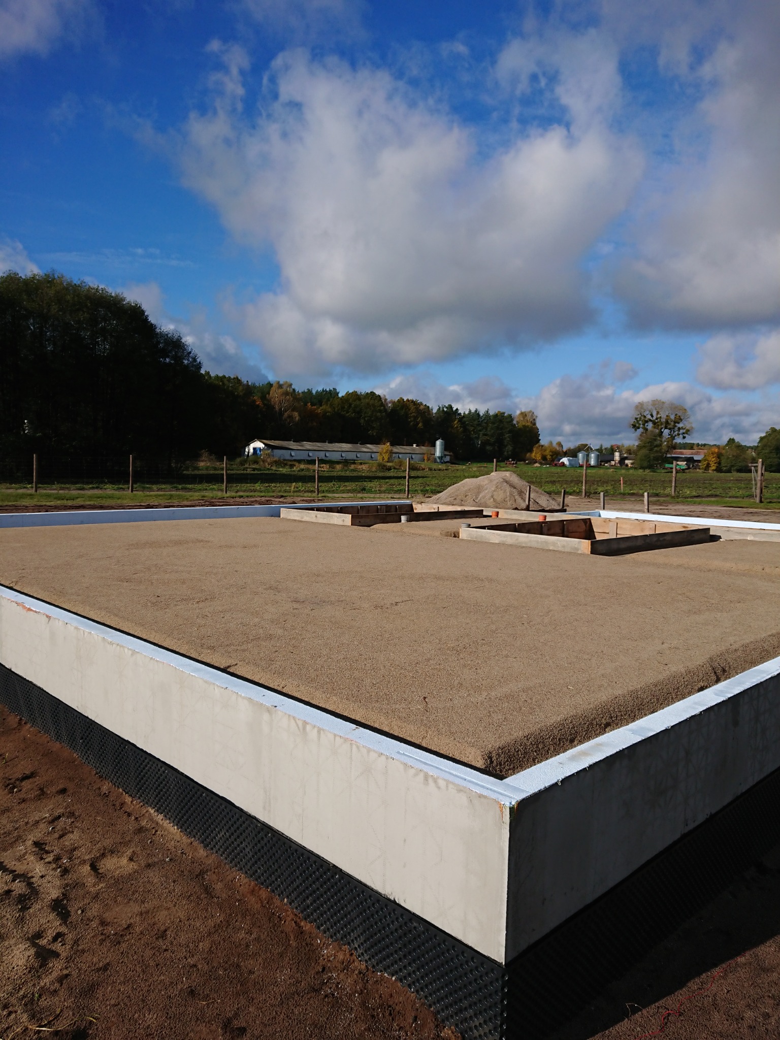 Przygotowany teren pod wylewkę fundamentów z widoczną hydroizolacją pionową oraz szalunkami pod przyszłe ściany nośne, w tle krajobraz wiejski.