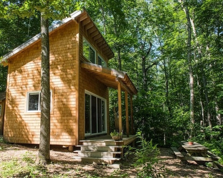 Drewniany domek letniskowy z elewacją pokrytą gontem, otoczony zielonym lasem, z widocznym tarasem wejściowym i schodami oraz stołem piknikowym obok.