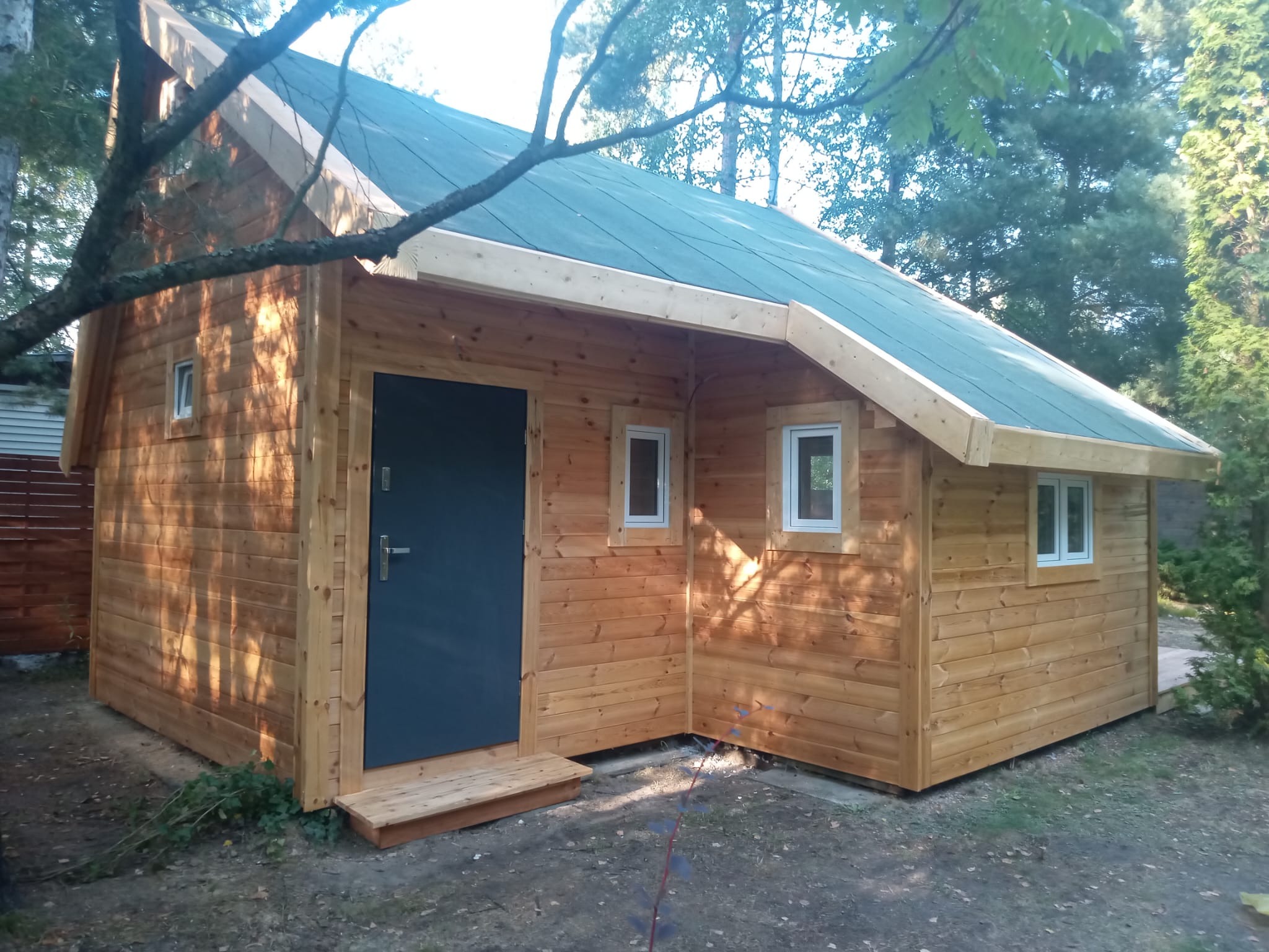 Nowoczesna, drewniana konstrukcja z płaskim, ciemnym dachem i szarą, metalową furtką wejściową, otoczona zielenią drzew i krzewów.