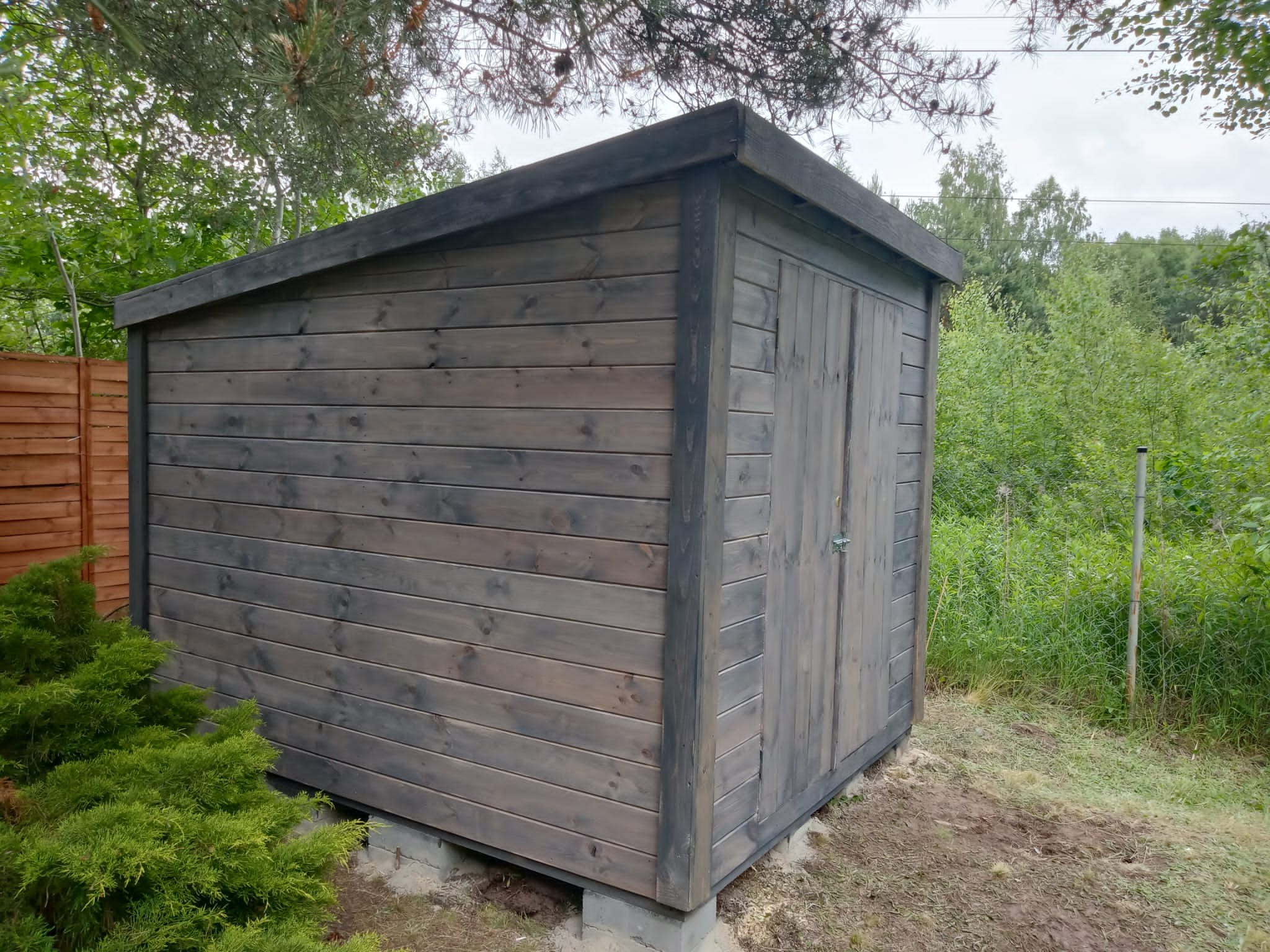 Drewniana szopa narzędziowa z dwuspadowym dachem, pomalowana na ciemnoszaro, osadzona na betonowych bloczkach, otoczona zielenią.