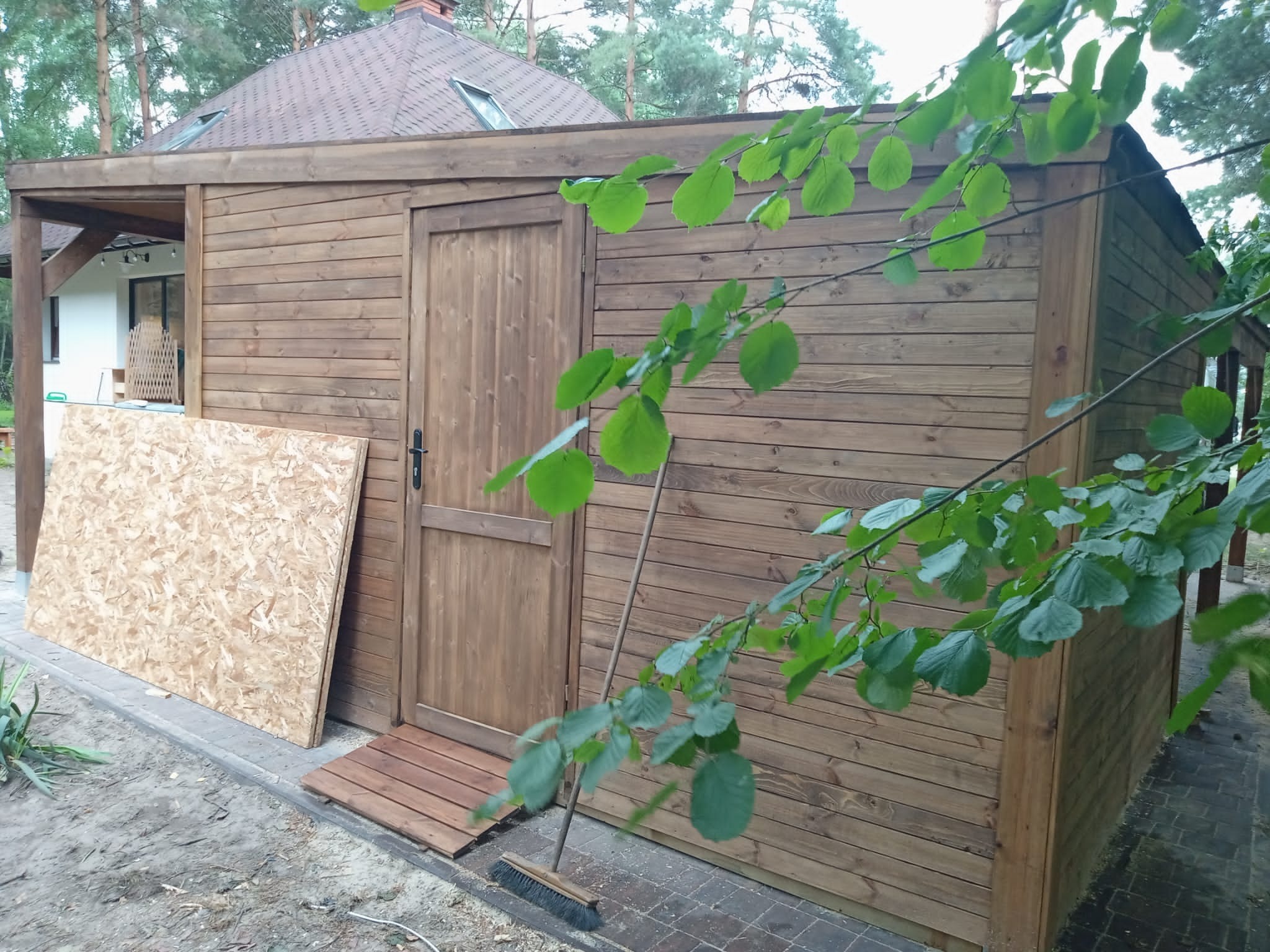 Brązowa, drewniana konstrukcja z drzwiami i fragmentem zadaszenia, obok leży płyta OSB, widoczny fragment domu w tle