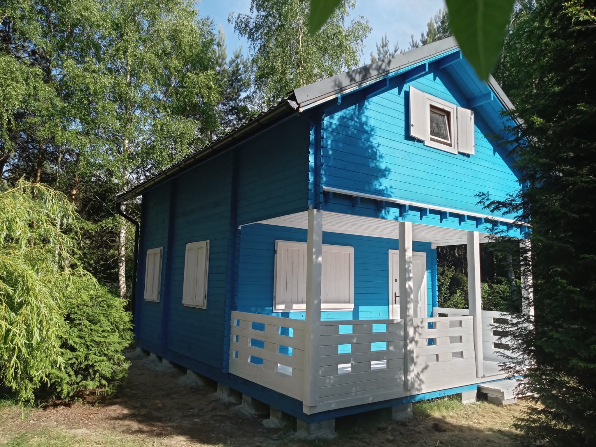 Niebieski domek letniskowy z białymi okiennicami i werandą, otoczony zielenią drzew i krzewów w słoneczny dzień.
