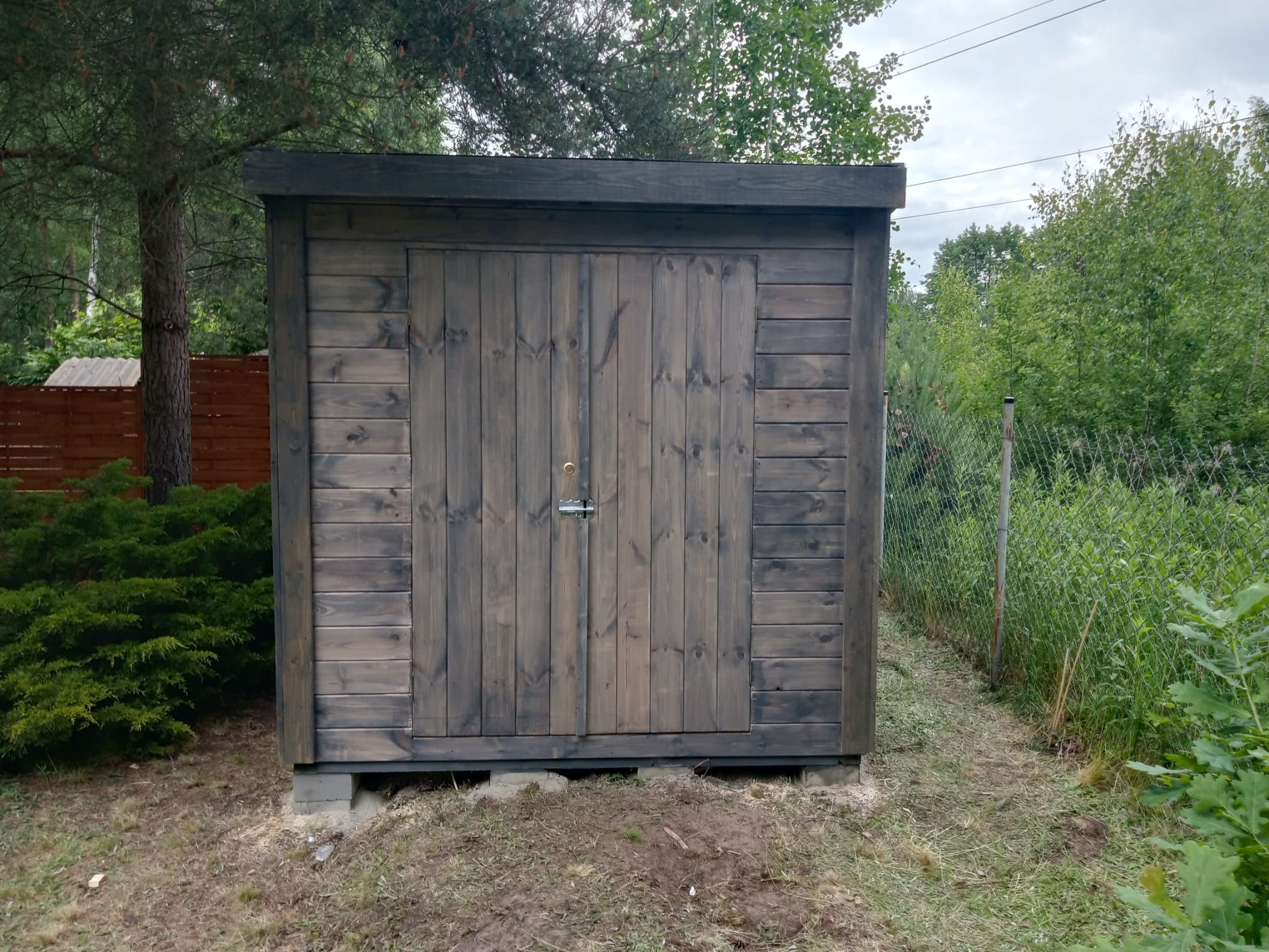 Drewniana konstrukcja z dwuskrzydłowymi drzwiami w ogrodzie, pomalowana na szaro, z metalowym zamkiem, osadzona na betonowych bloczkach, otoczona zielenią.