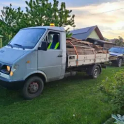 Szara furgonetka z drewnem na pace, zaparkowana na trawniku przed domem, w tle widoczny samochód terenowy z przyczepką.