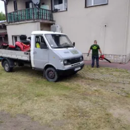 Pracownik w zielonej koszulce z logo firmy, stojący obok dostawczego samochodu z czerwoną kosiarką na pace i dmuchawą do liści w rękach, na tle domu z balkonem.