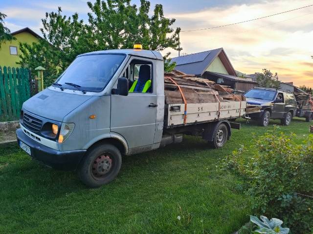 Szara furgonetka z drewnem na pace, zaparkowana na trawniku przed domem, w tle widoczny samochód terenowy z przyczepką.