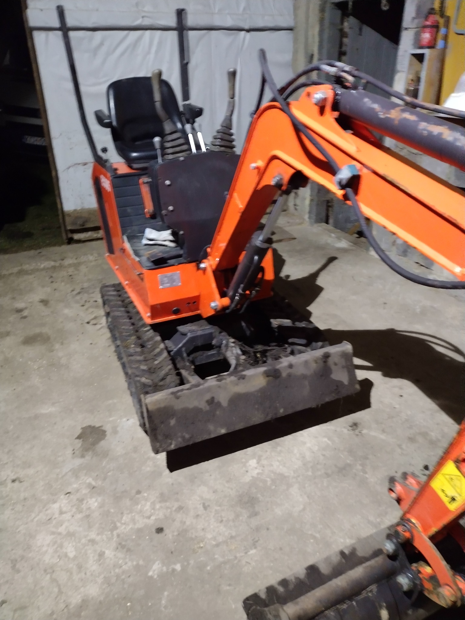 Pomarańczowa mini koparka gąsienicowa z łyżką, widoczna z boku, na betonowej posadzce w słabo oświetlonym pomieszczeniu.