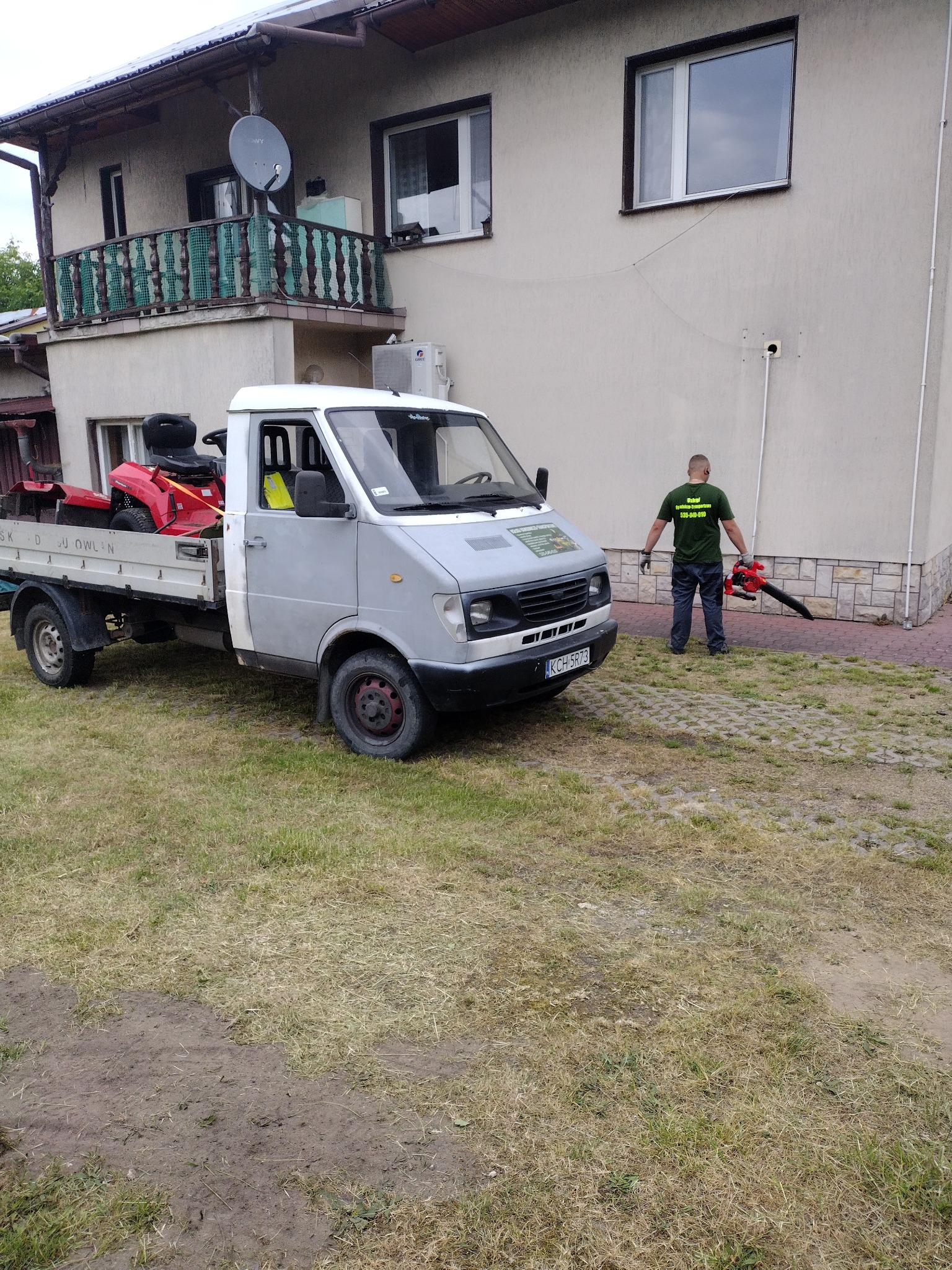 Pracownik w zielonej koszulce z logo firmy, stojący obok dostawczego samochodu z czerwoną kosiarką na pace i dmuchawą do liści w rękach, na tle domu z balkonem.