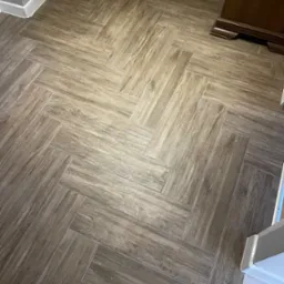 Ułożona w jodełkę podłoga z płytek imitujących drewno, z widoczną listwą przypodłogową i fragmentem ciemnych mebli.