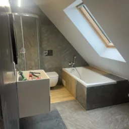 Ukończona łazienka na poddaszu ze skośnym oknem dachowym, szarą płytką imitującą kamień na podłodze i ścianach, białą wanną obudowaną drewnem i szarym kamieniem, oraz wiszącą szafką z umywalką,...