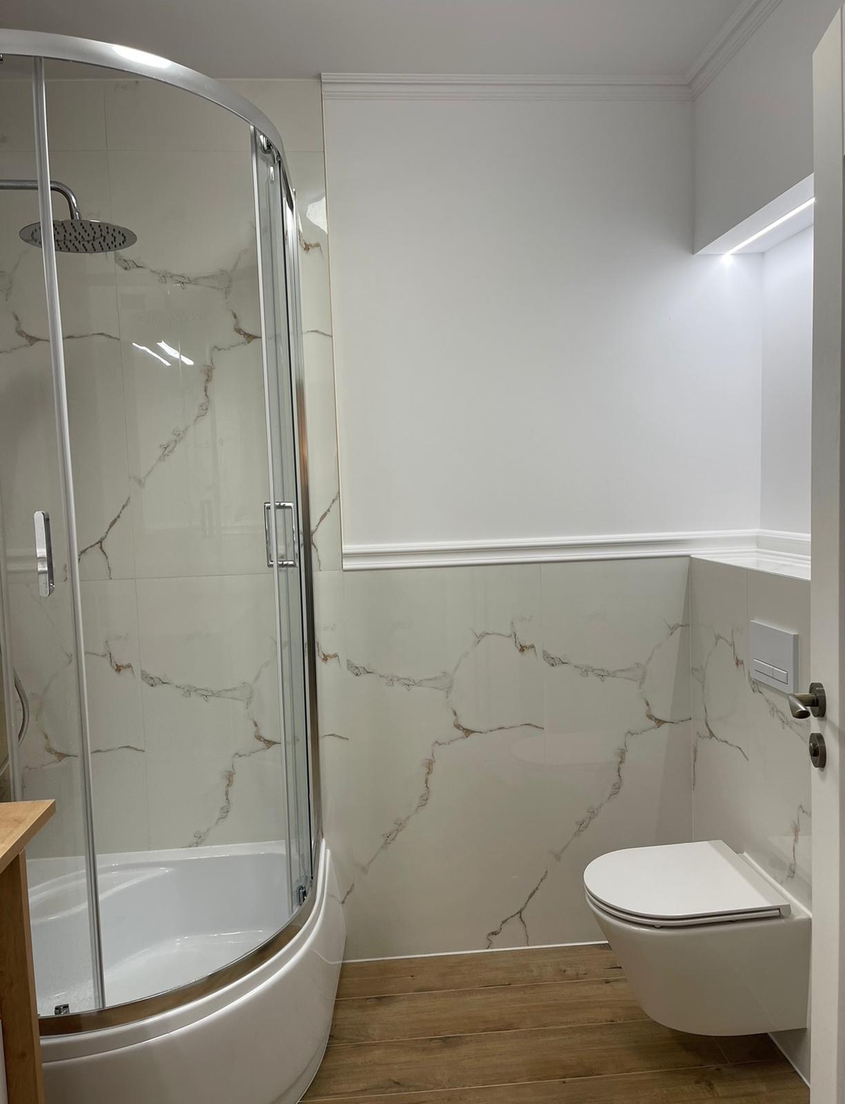 Nowoczesna łazienka z kabiną prysznicową z zaokrąglonym szkłem, białą ceramiką sanitarną i płytkami imitującymi marmur z brązowymi żyłami na ścianach, drewnopodobna podłoga i dekoracyjne listwy...