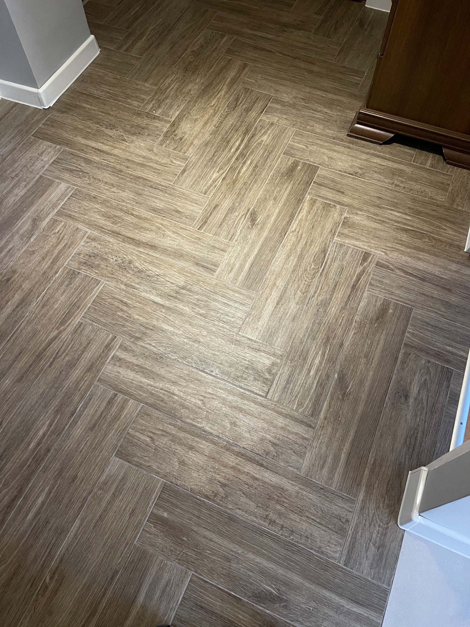 Ułożona w jodełkę podłoga z płytek imitujących drewno, z widoczną listwą przypodłogową i fragmentem ciemnych mebli.