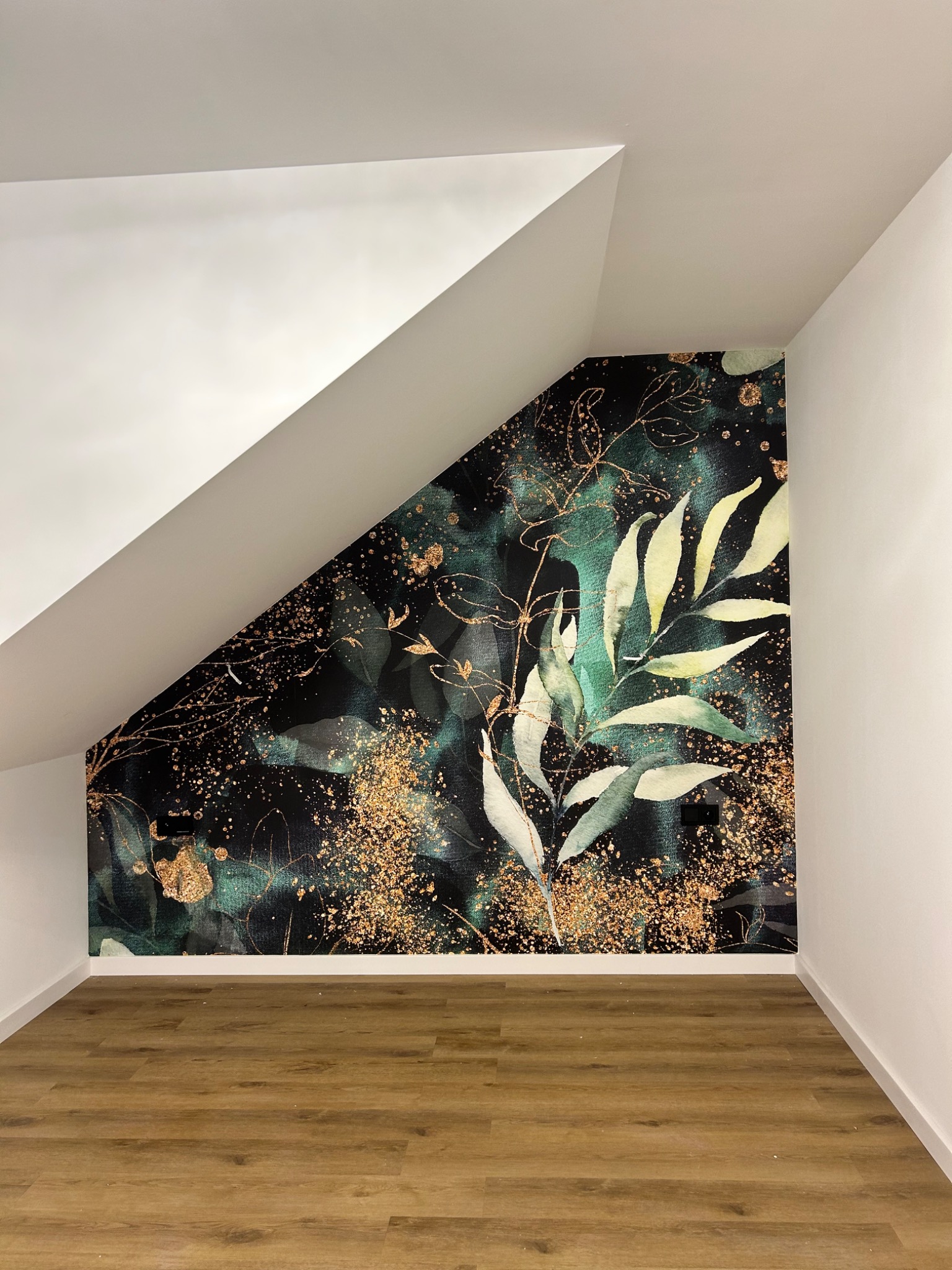 Pokój na poddaszu z drewnianą podłogą, białe ściany i dekoracyjna tapeta z motywem liści i złotych kropek na ścianie szczytowej.