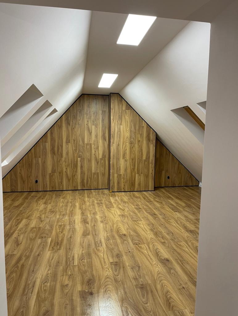 Wykończone poddasze z drewnopodobną podłogą i zabudową wnęki pod skosem, oświetlone dwoma lampami sufitowymi.