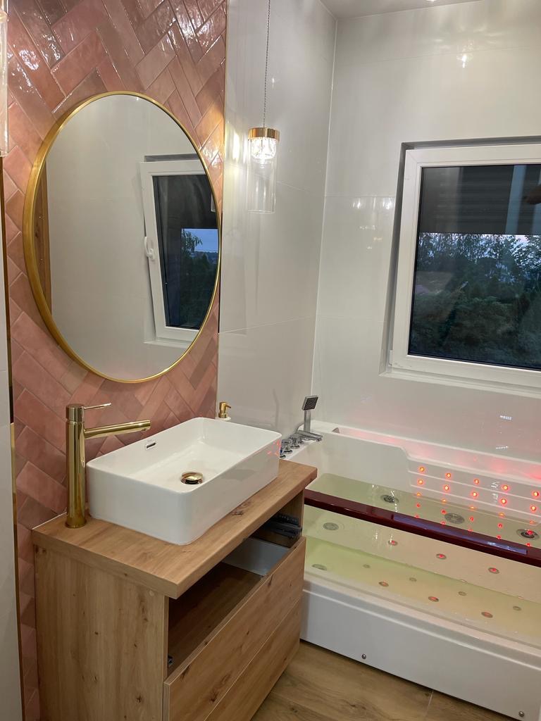Nowoczesna łazienka z umywalką nablatową na drewnianej szafce, okrągłe lustro w złotej ramie na tle ściany wyłożonej różowymi płytkami w jodełkę, wanna z hydromasażem i oświetleniem LED.