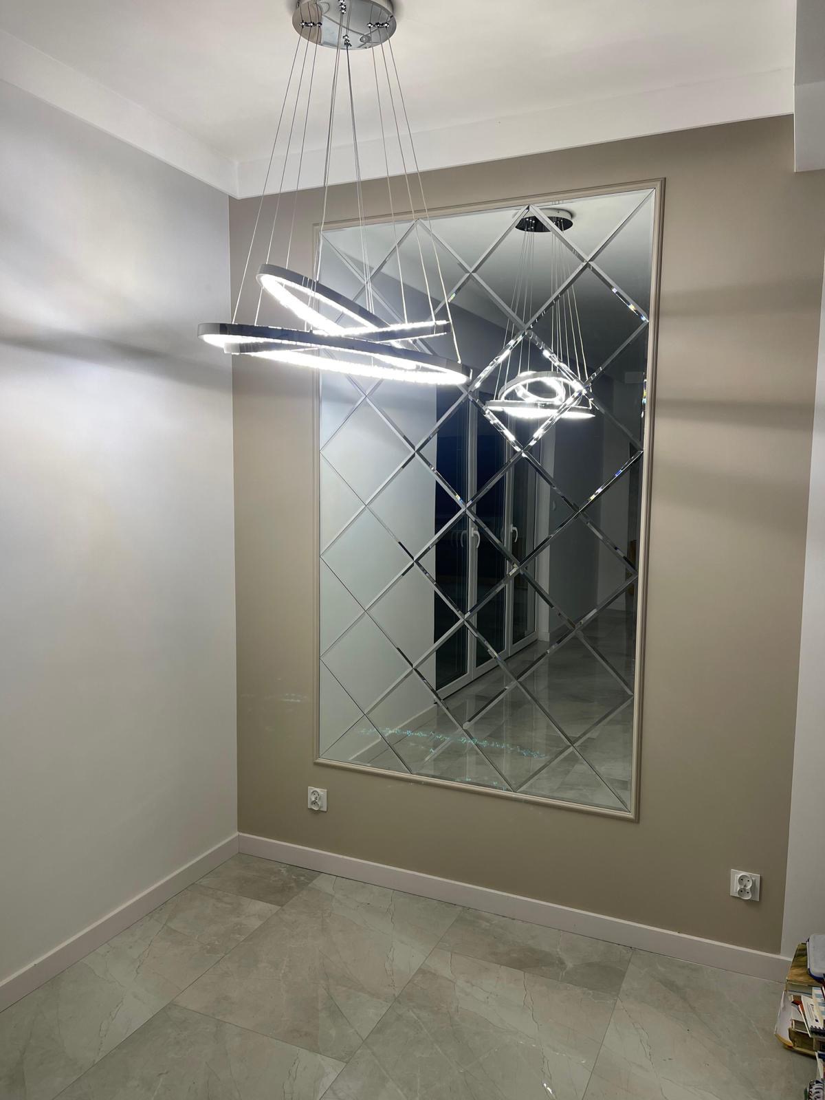 Lustrzana ściana z romboidalnych elementów, oświetlona nowoczesną lampą wiszącą z pierścieni LED, odbijająca widok wnętrza z oknem balkonowym.