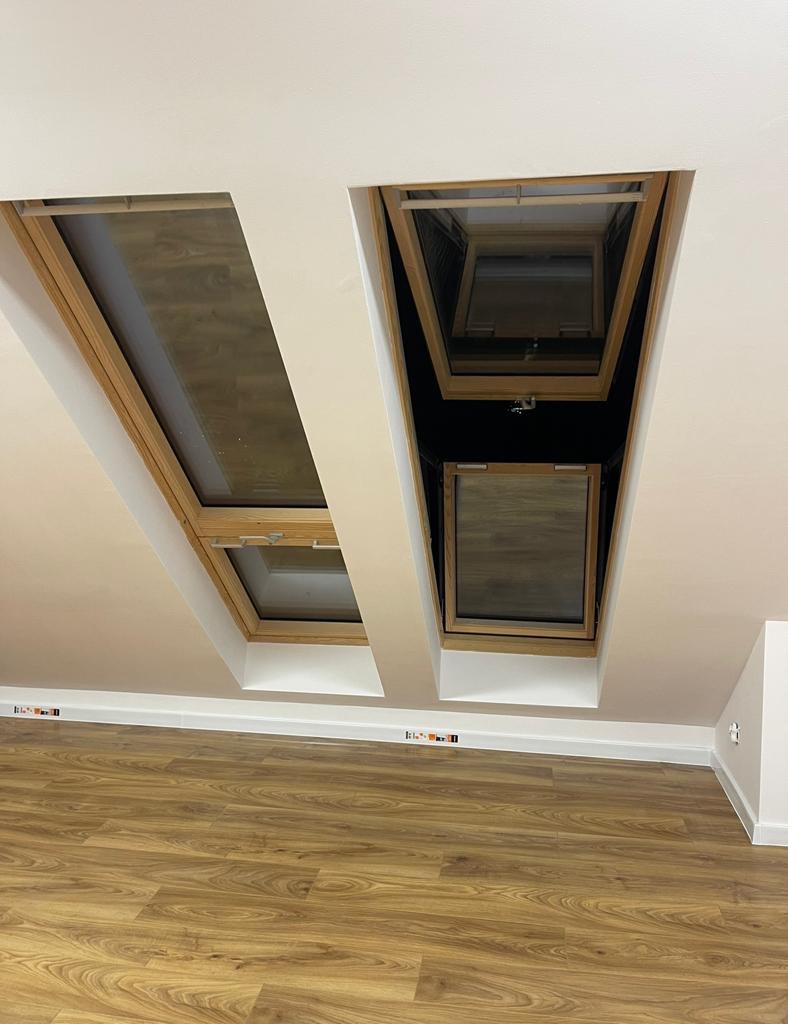Dwa okna dachowe z drewnianymi ramami, zamontowane w białym suficie pod skosem, widoczne z perspektywy podłogi z paneli imitujących drewno.
