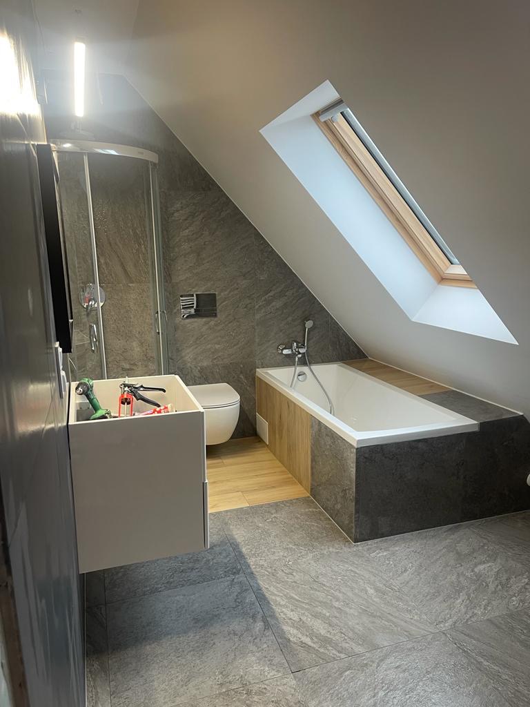 Ukończona łazienka na poddaszu ze skośnym oknem dachowym, szarą płytką imitującą kamień na podłodze i ścianach, białą wanną obudowaną drewnem i szarym kamieniem, oraz wiszącą szafką z umywalką,...