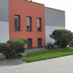 Nowoczesny ogród przydomowy z równo przystrzyżonymi krzewami, kamieniami ozdobnymi i fragmentem trawnika, na tle budynku z elementami w kolorze łososiowym i szarym.