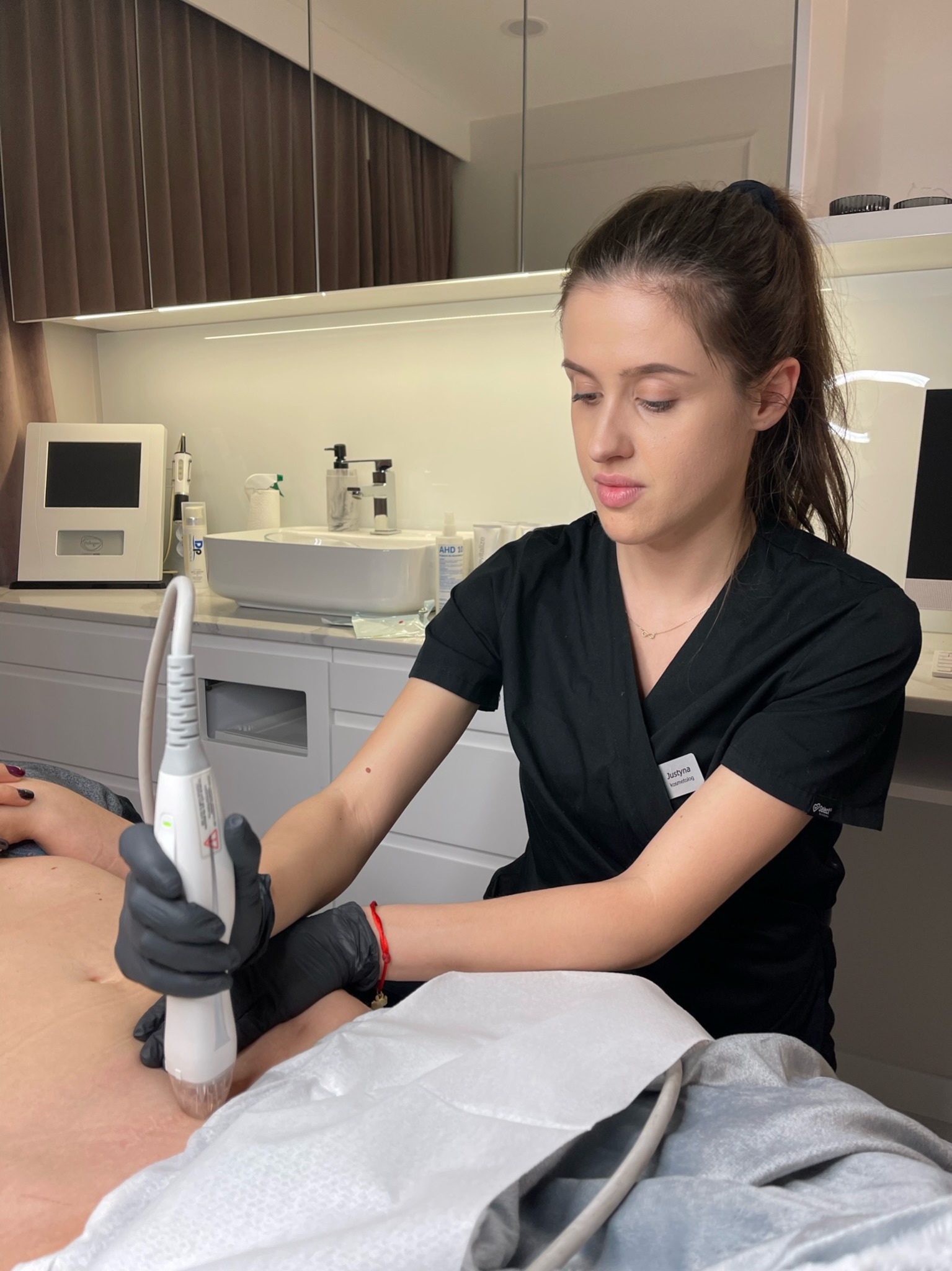 Kosmetolog w czarnym uniformie, w czarnych rękawiczkach wykonuje zabieg na brzuchu pacjenta przy użyciu urządzenia z białą głowicą w sterylnym gabinecie.