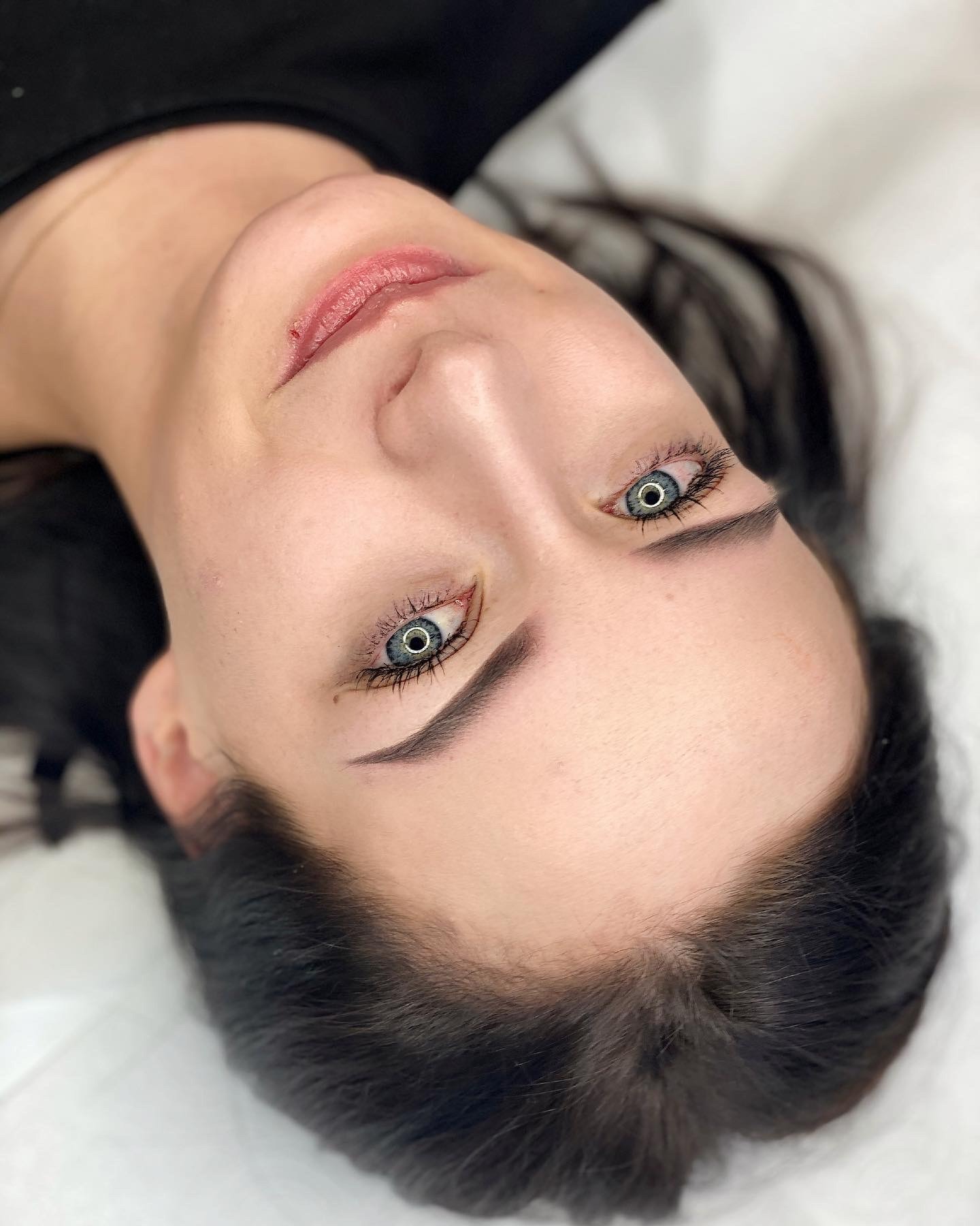 Kobieta z niebieskimi oczami leżąca na białym podłożu, po zabiegu pigmentacji brwi metodą microblading, widoczne drobne zaczerwienienia wokół ust.