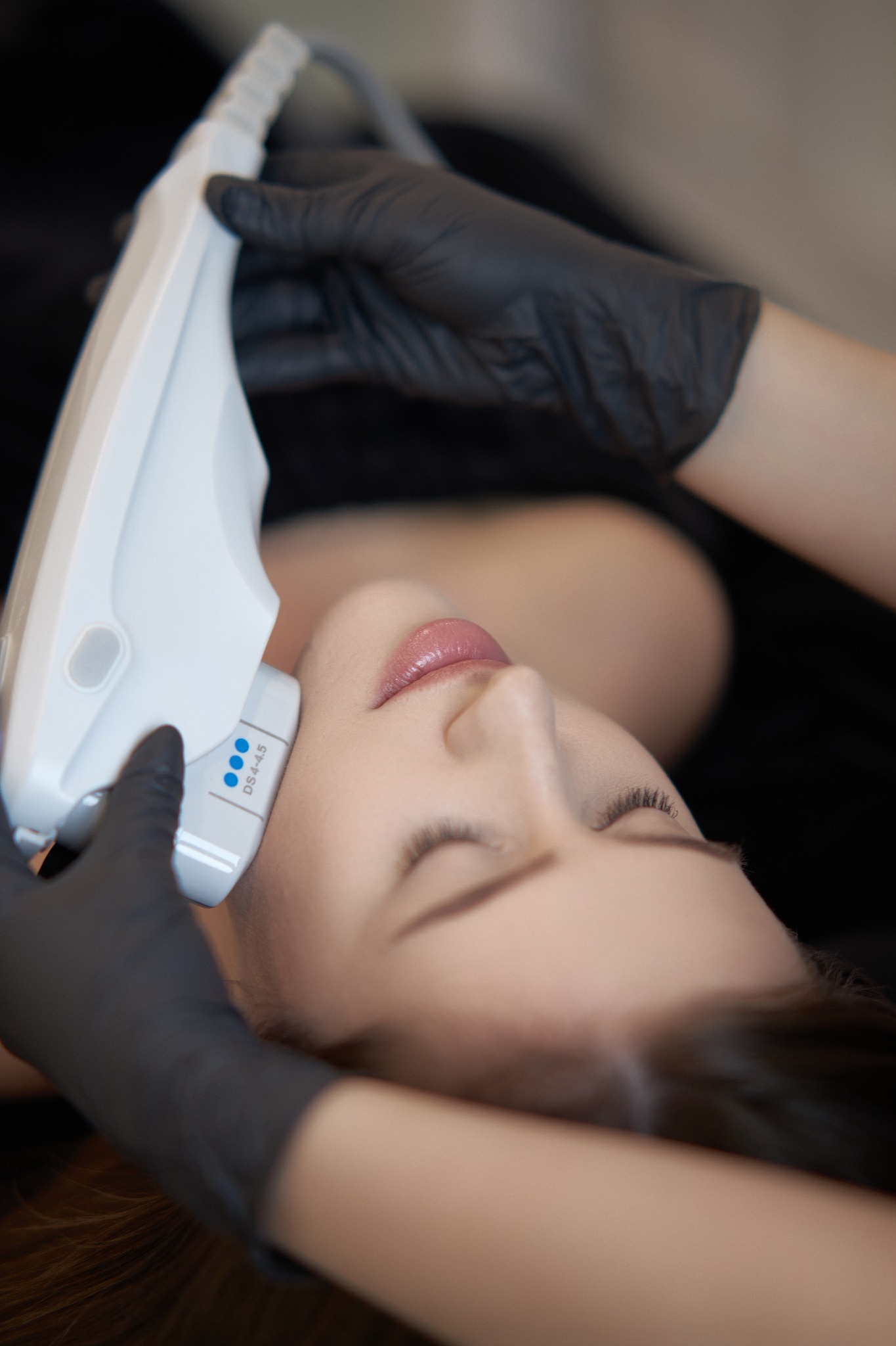 Zabieg HIFU na twarz pacjentki, widoczny aplikator z oznaczeniem DS4-4.5, dłonie w czarnych rękawiczkach, zamknięte oczy i delikatny makijaż.