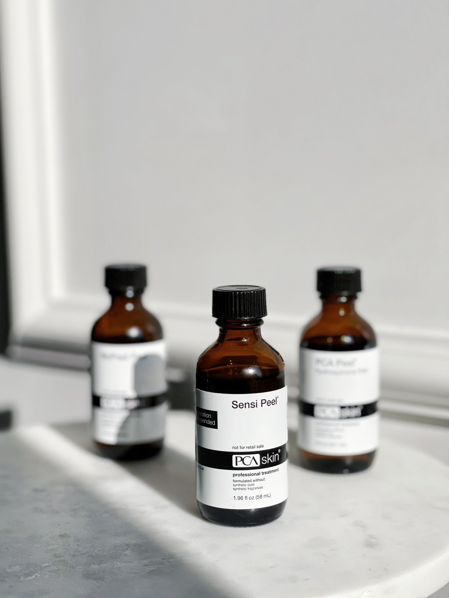 Trzy brązowe butelki PCA Skin Sensi Peel z czarnymi nakrętkami, ustawione na marmurowej powierzchni, z białą ścianą w tle.