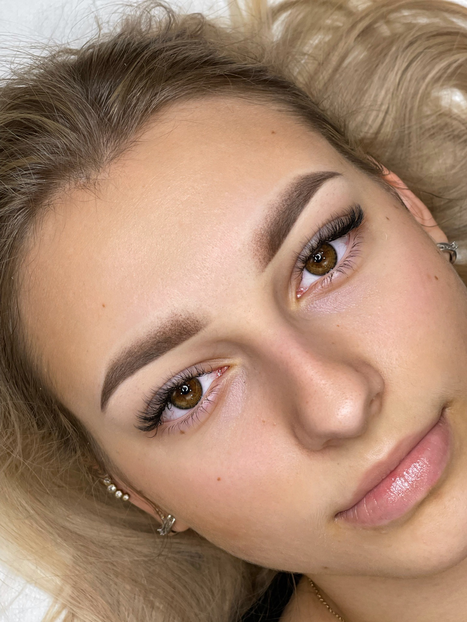 Zbliżenie na twarz kobiety po zabiegu laminacji brwi i przedłużania rzęs, widoczne delikatne kolczyki.