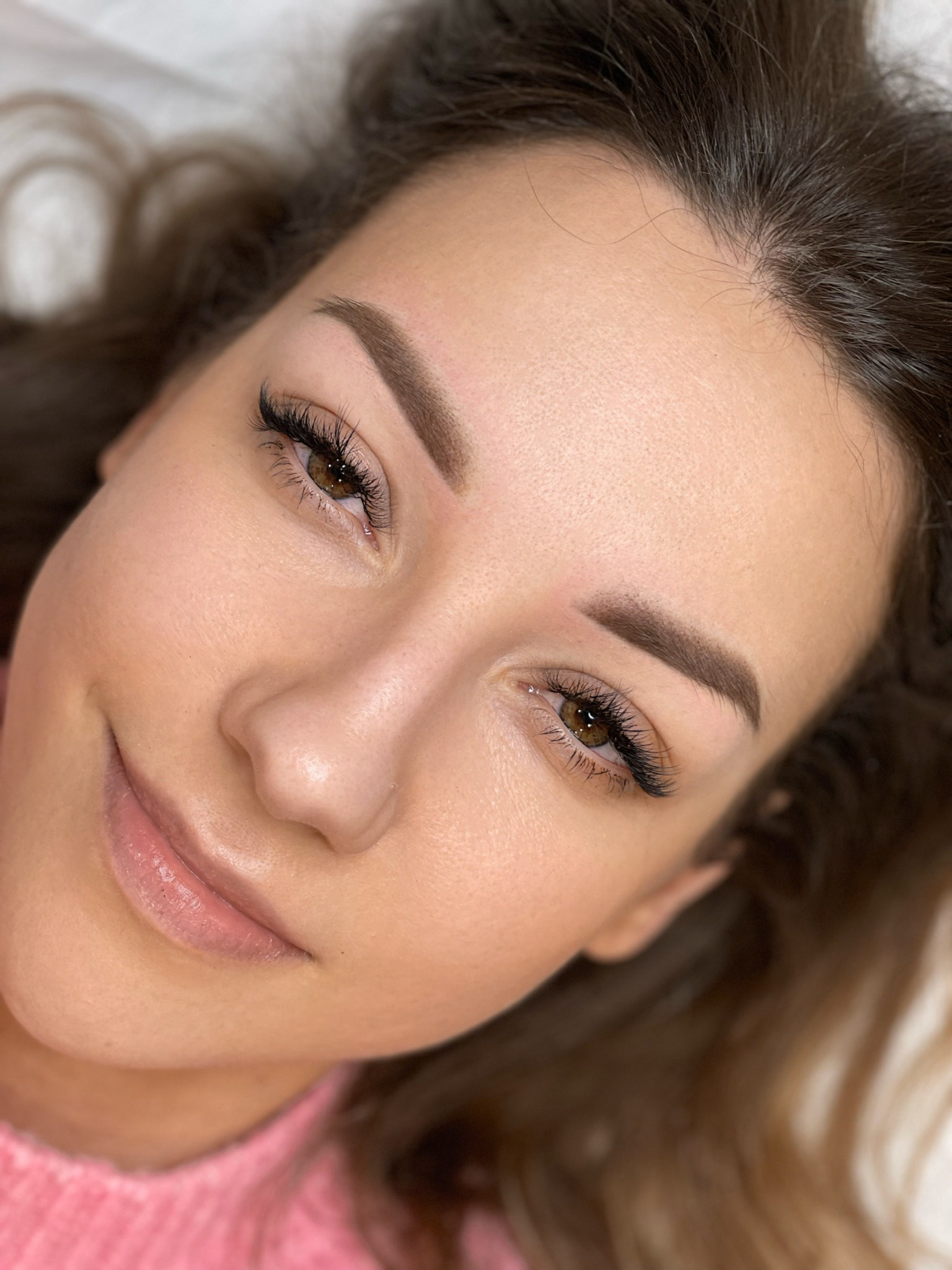 Portret kobiety z delikatnym uśmiechem, prezentującej brwi po zabiegu microbladingu i przedłużone rzęsy, z widocznymi drobnymi niedoskonałościami skóry.